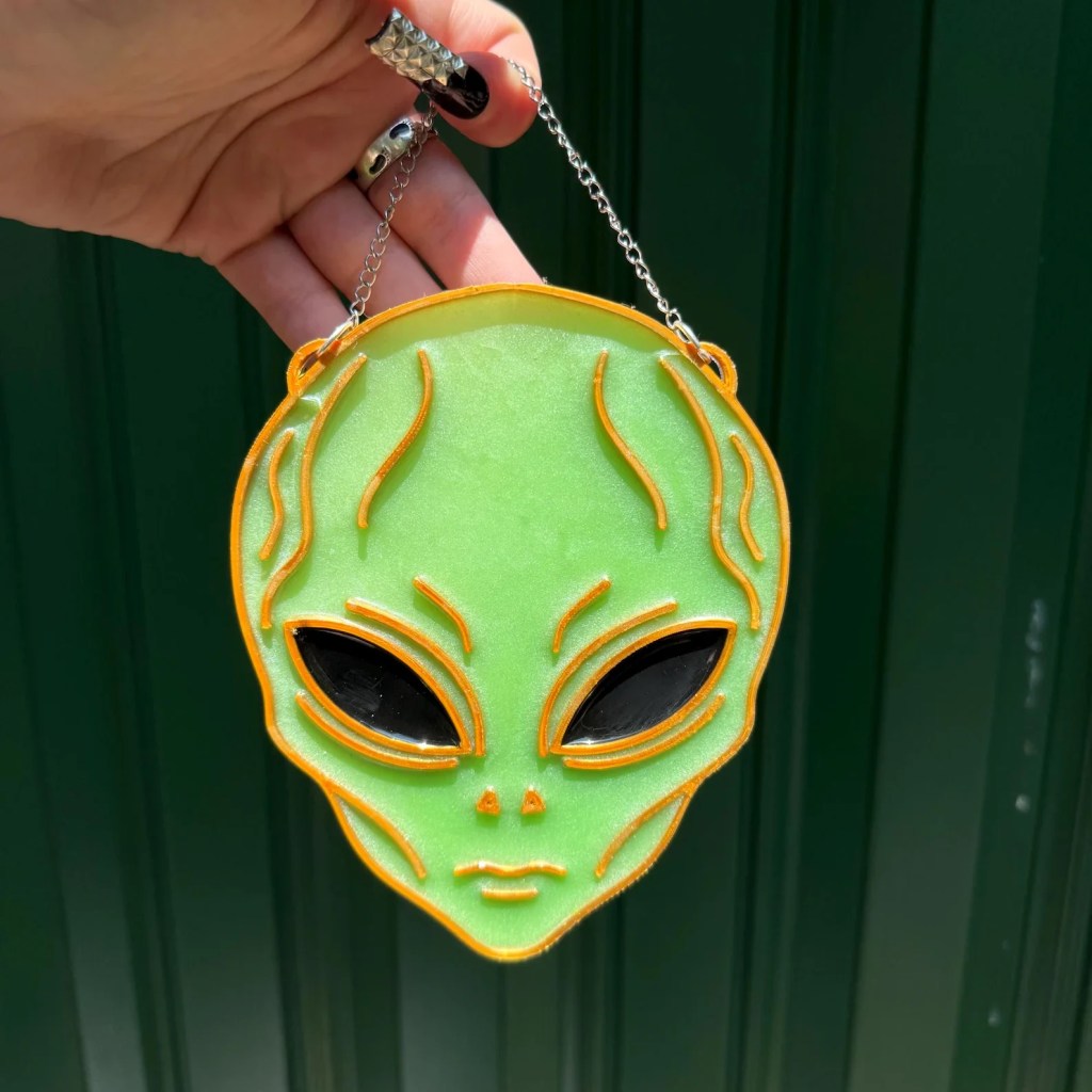 A green alien head pendant with orange outlining and black eyes, suspended by a silver chain. 